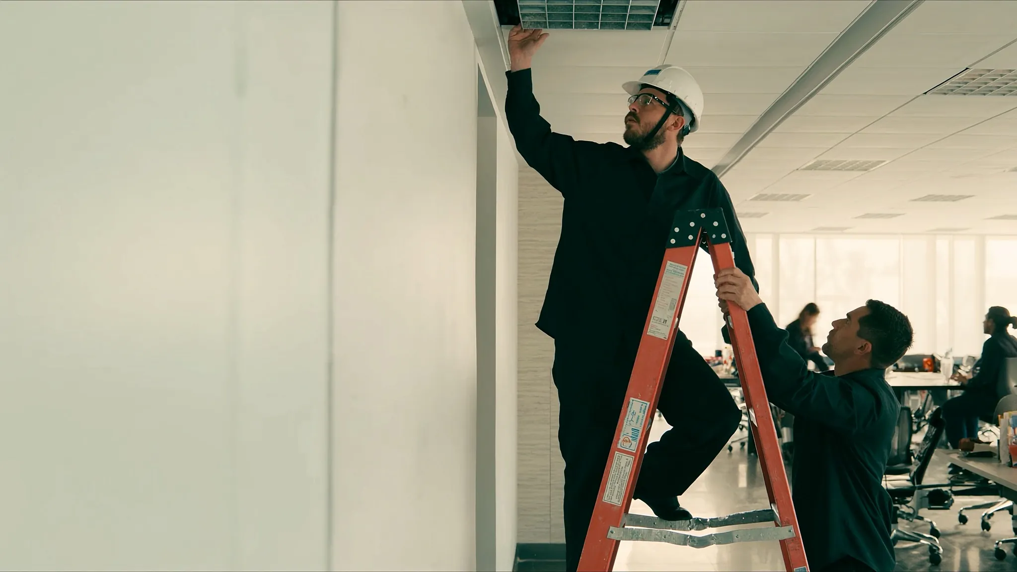 Técnico cambia una luminaria sobre una escalera de fibra de vidrio mientras un compañero la asegura en una oficina